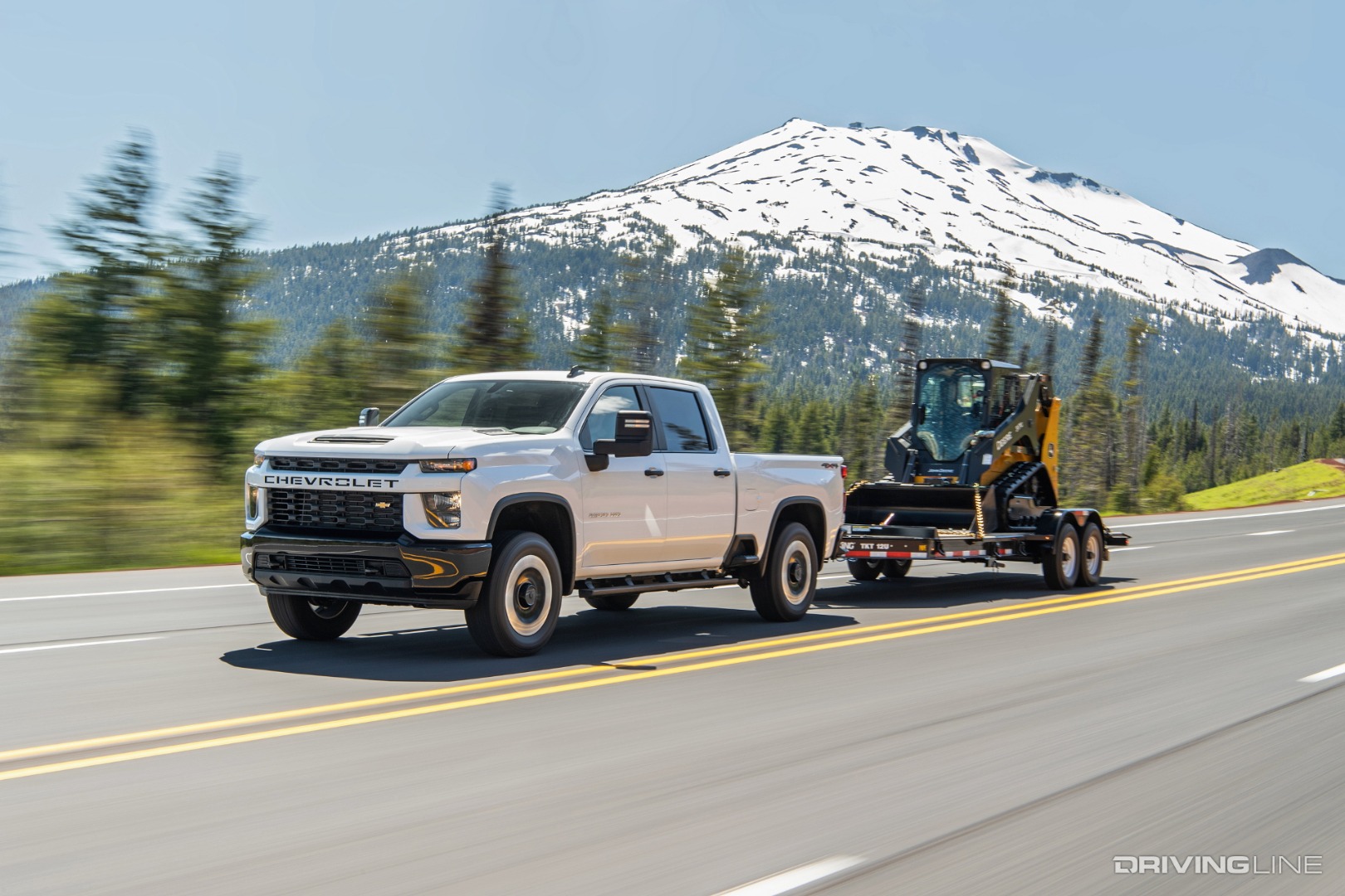 2021 Silverado pulling a trailer with a skid loader on it down a road.